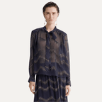 A model wearing a sheer long-sleeve button-up blouse with a tie collar and a matching skirt, both featuring a dark navy blue and beige wavy abstract pattern, set against a plain light gray background.