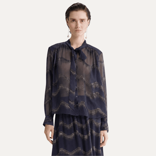 A model wearing a sheer long-sleeve button-up blouse with a tie collar and a matching skirt, both featuring a dark navy blue and beige wavy abstract pattern, set against a plain light gray background.