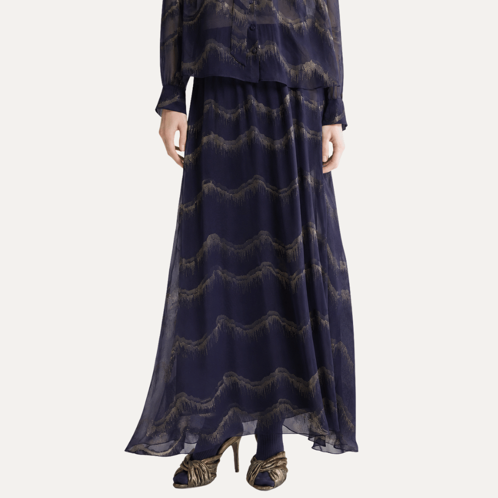 A navy blue two-piece outfit featuring a long-sleeve sheer top and a matching maxi skirt with a wavy metallic gold pattern, shown on a model against a light gray background.