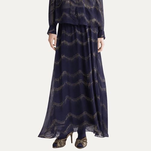 A navy blue two-piece outfit featuring a long-sleeve sheer top and a matching maxi skirt with a wavy metallic gold pattern, shown on a model against a light gray background.