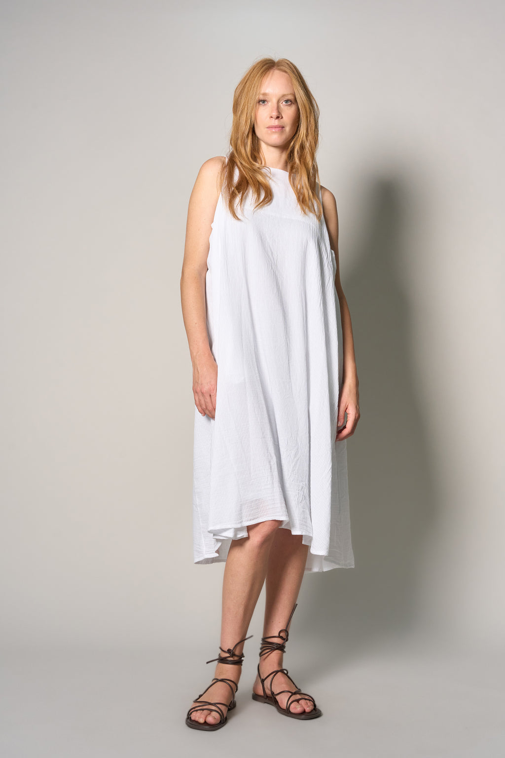 A woman modeling a sleeveless white high-low dress with a loose, flowing silhouette and black lace-up strappy sandals, standing against a neutral light gray studio background.