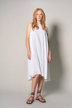 A woman modeling a sleeveless white high-low dress with a loose, flowing silhouette and black lace-up strappy sandals, standing against a neutral light gray studio background.