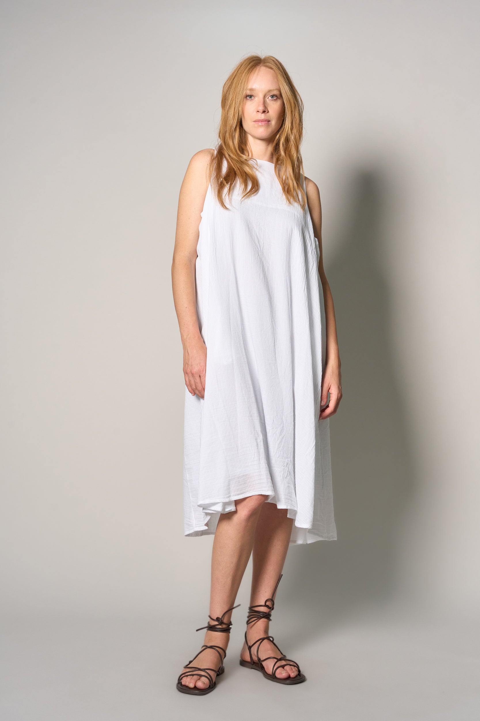 A woman modeling a sleeveless white high-low dress with a loose, flowing silhouette and black lace-up strappy sandals, standing against a neutral light gray studio background.