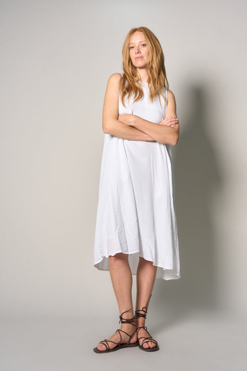A woman with blonde hair stands in a studio wearing a sleeveless white midi dress and black strappy flat sandals against a light gray background.
