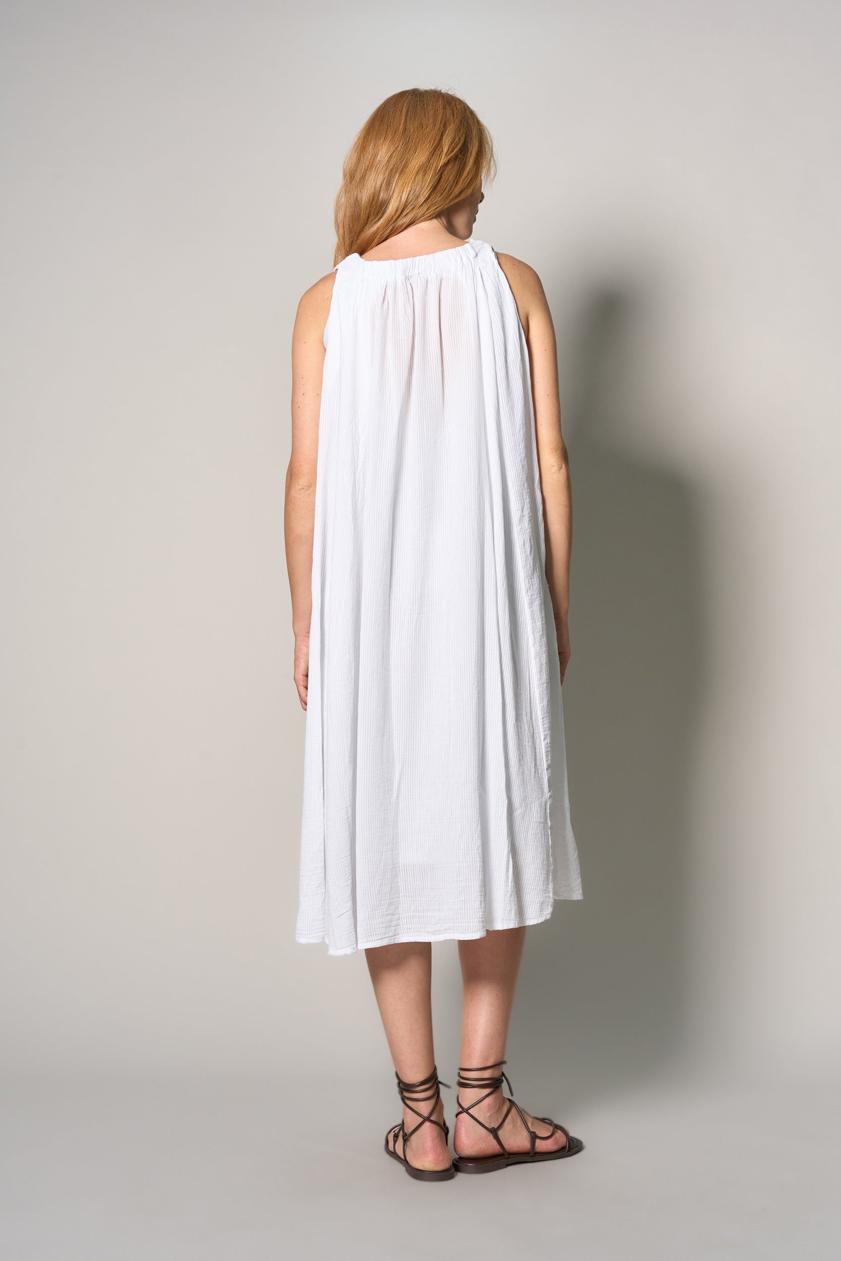 A woman viewed from behind wearing a sleeveless white midi dress with a gathered neckline and black lace-up flat sandals, standing against a light gray studio background.