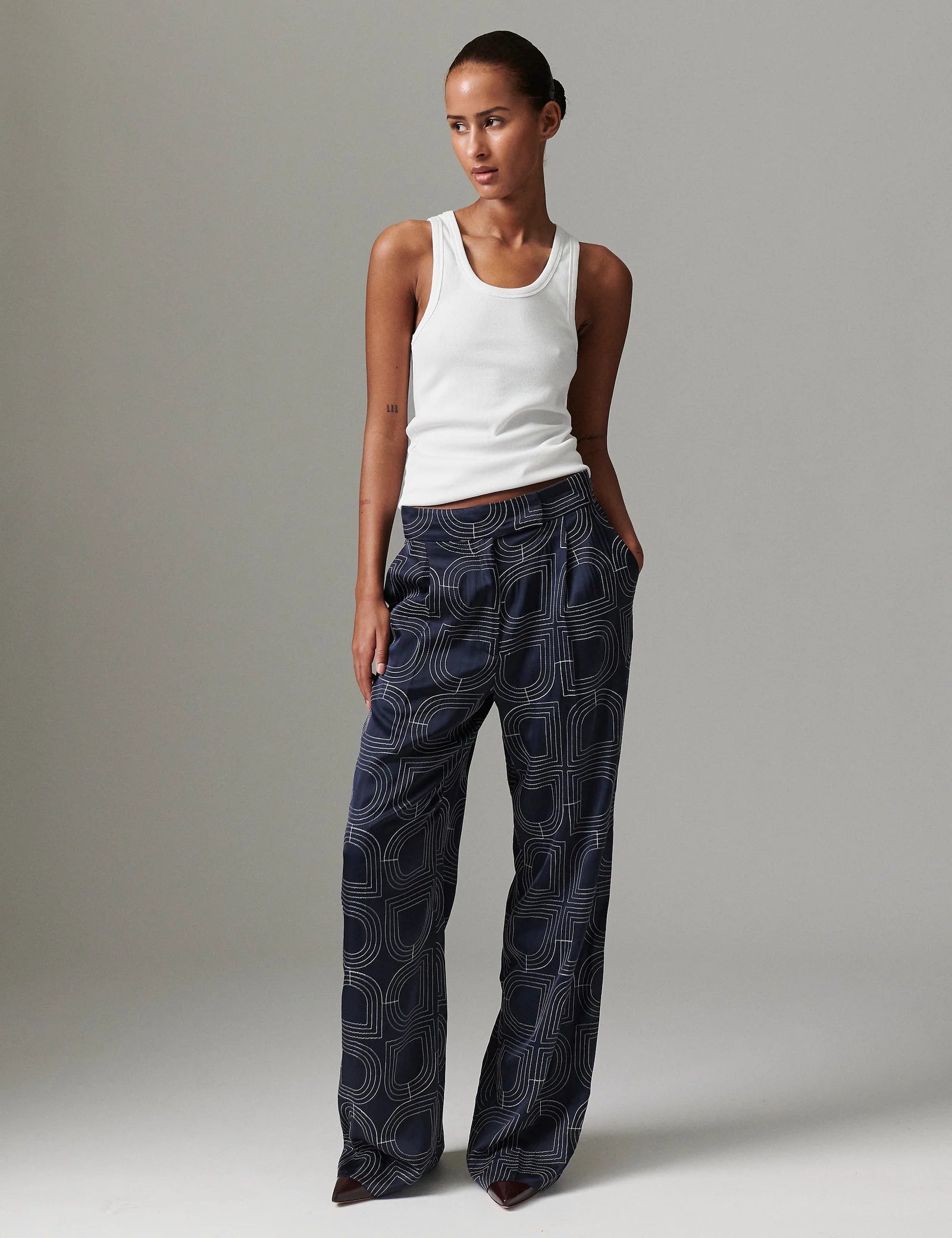 Woman wearing a white tank top and dark patterned pants on a plain background
