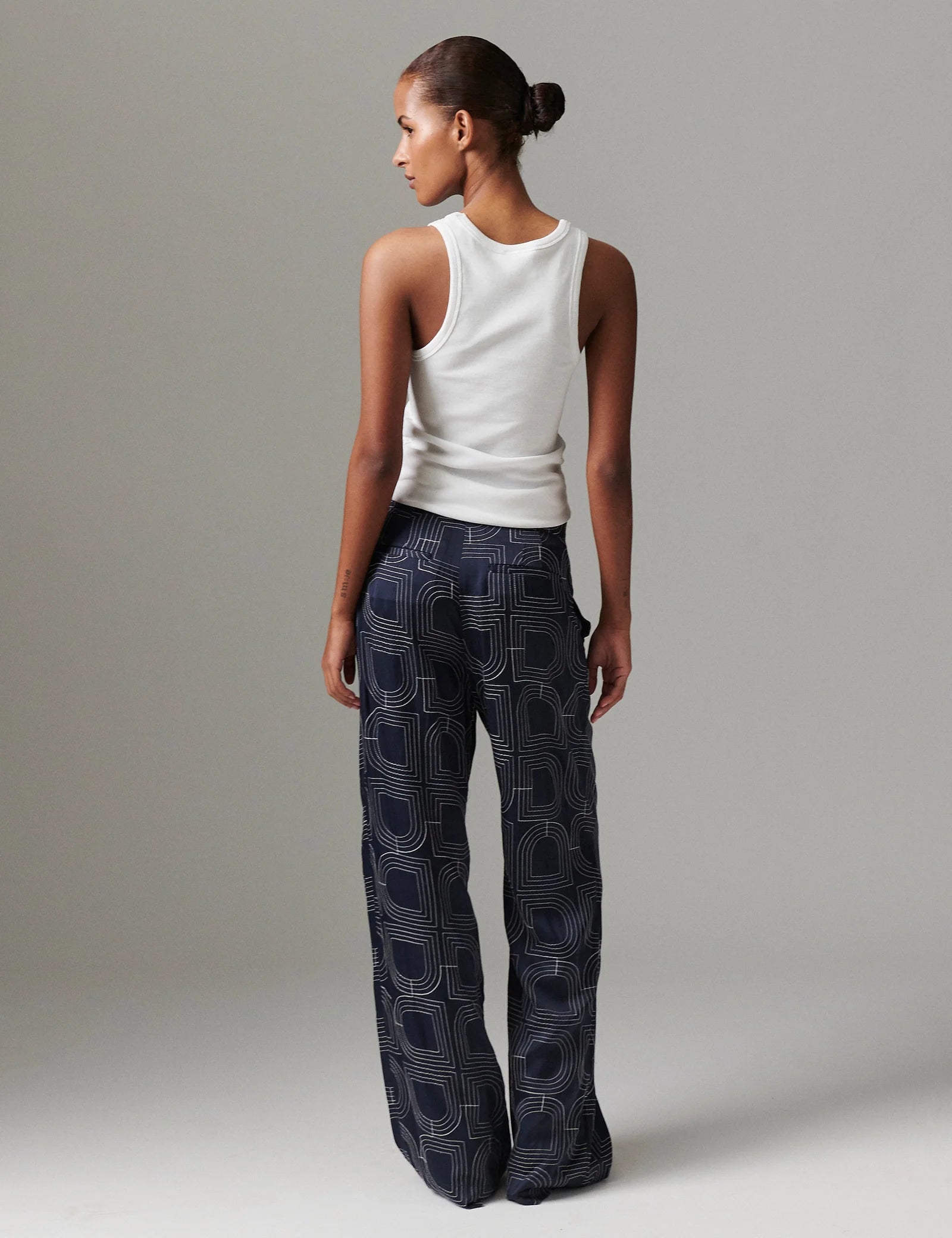 Woman wearing a white tank top and dark patterned pants on a plain background