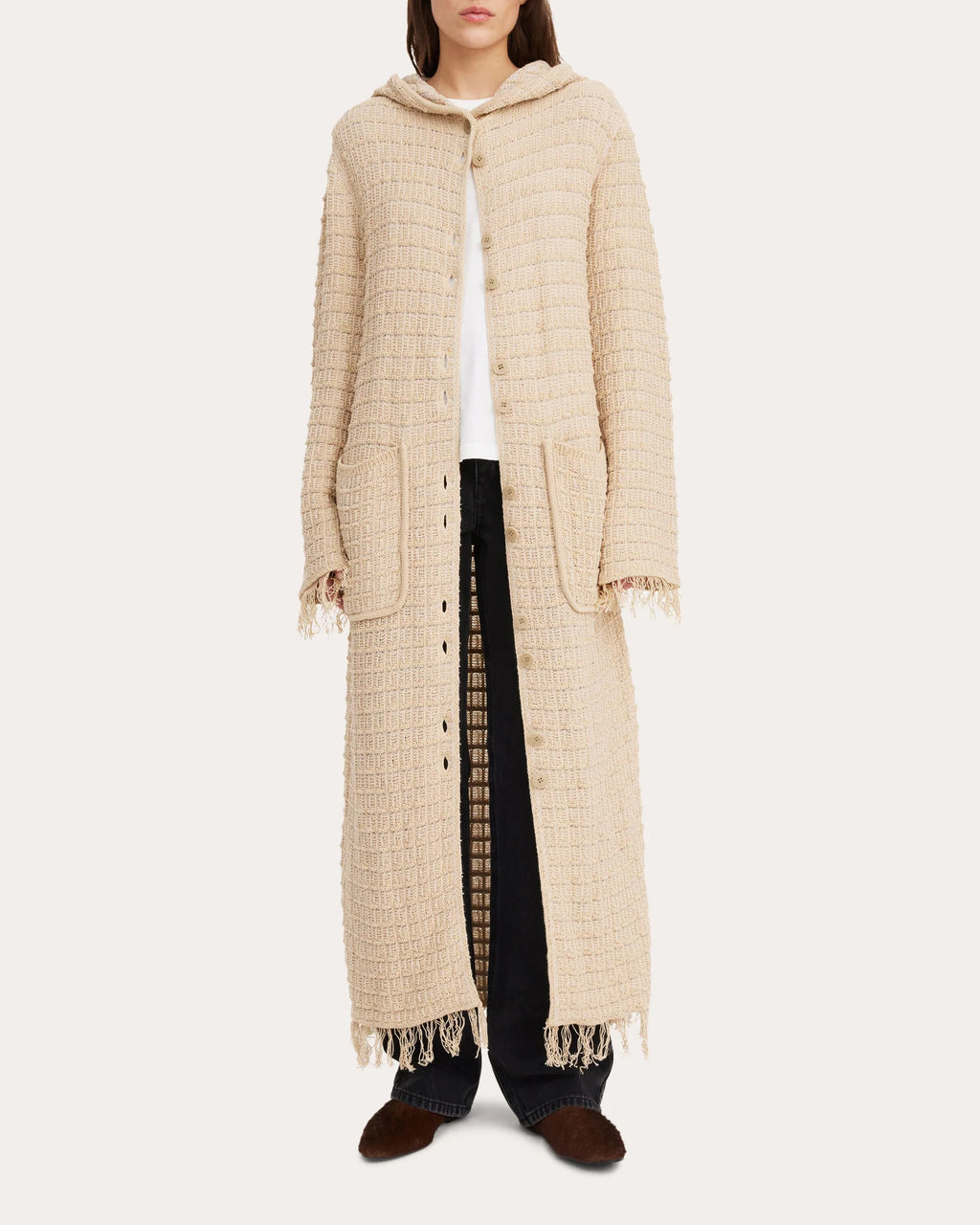A model wearing a beige knitted fringe jacket with a hood, paired with black trousers.
