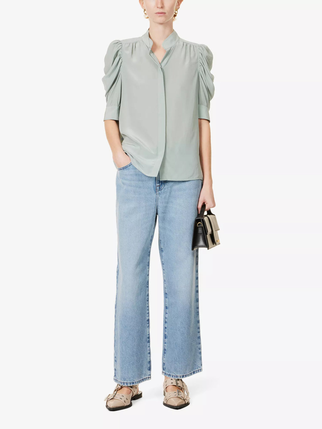 A woman wearing a high-necked, short-sleeved green silk blouse with puff sleeves, tucked into wide-leg blue jeans, with a slim belt and brown leather slides.