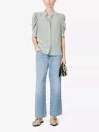 A woman wearing a high-necked, short-sleeved green silk blouse with puff sleeves, tucked into wide-leg blue jeans, with a slim belt and brown leather slides.