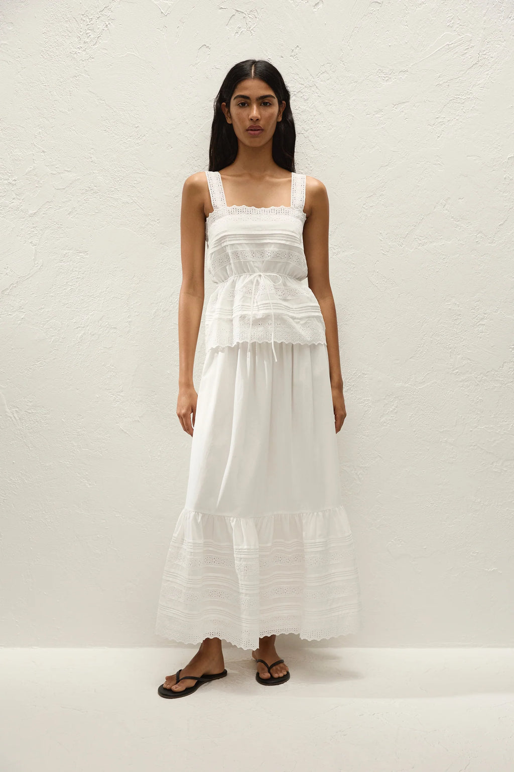 A woman modeling a white tiered maxi dress with ruffle details and a smocked bodice, standing against a textured white wall.