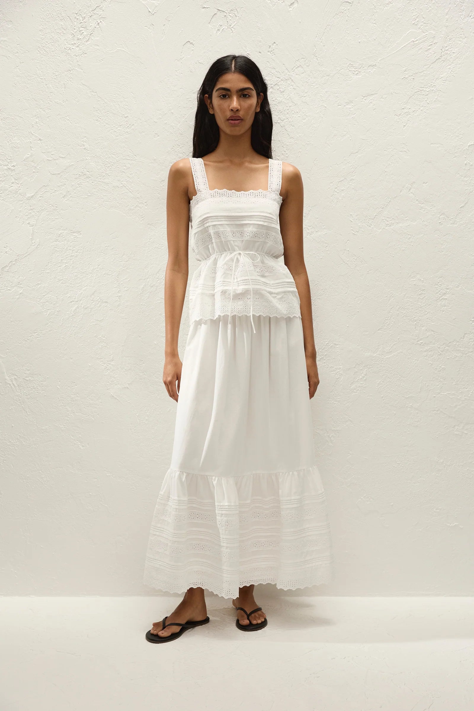 A woman modeling a white tiered maxi dress with ruffle details and a smocked bodice, standing against a textured white wall.