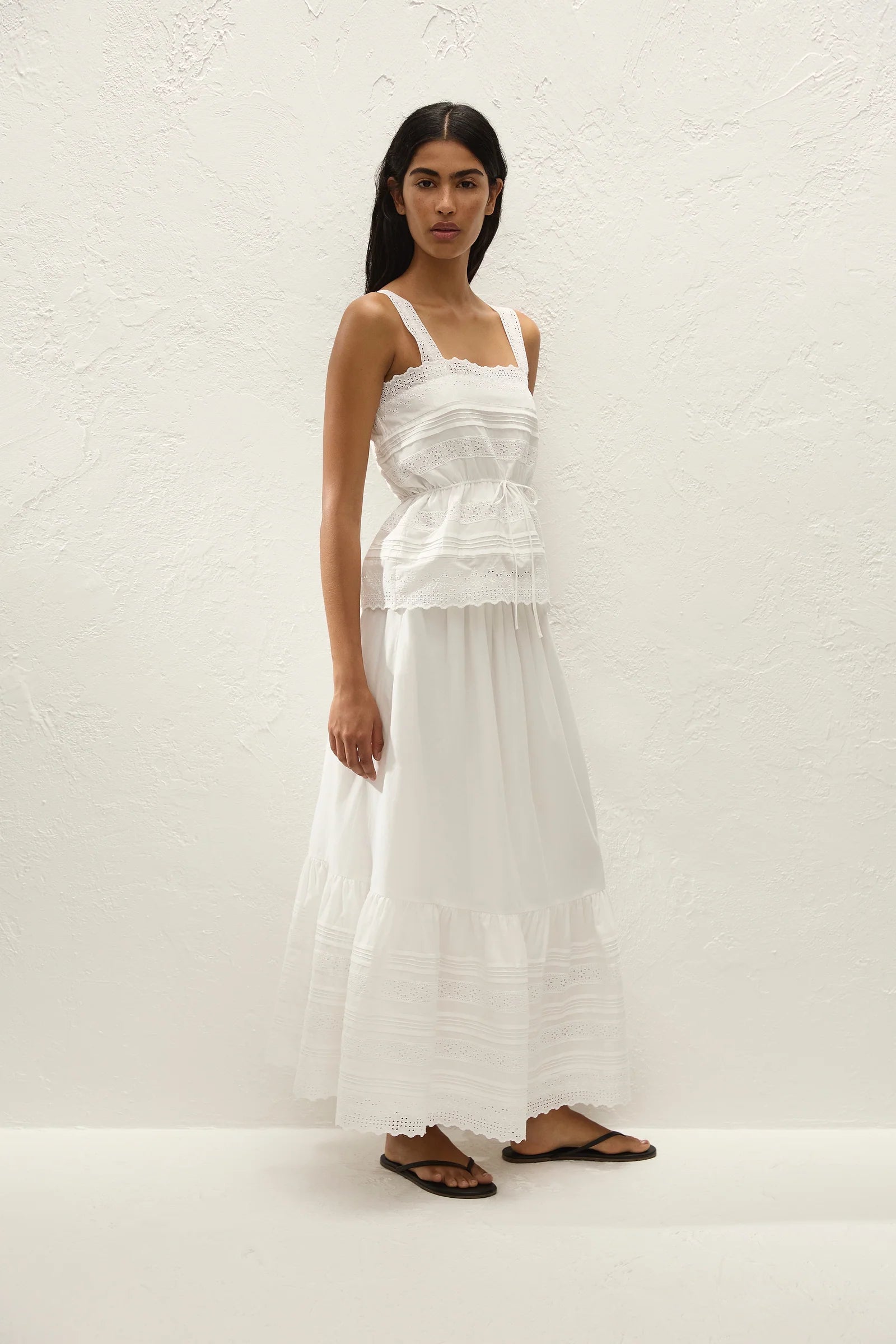 A woman modeling a white tiered maxi dress with thin shoulder straps and a gathered waist, standing against a textured off-white wall.