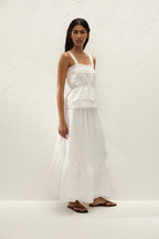A woman modeling a white tiered maxi dress with thin shoulder straps and a gathered waist, standing against a textured off-white wall.