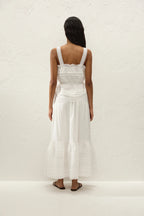 A woman viewed from behind wearing a white sleeveless maxi dress with a ruffled bodice and tiered skirt, standing against a textured off-white wall.