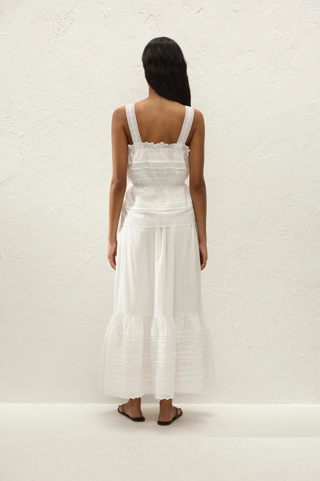 A woman viewed from behind wearing a white sleeveless maxi dress with a ruffled bodice and tiered skirt, standing against a textured off-white wall.