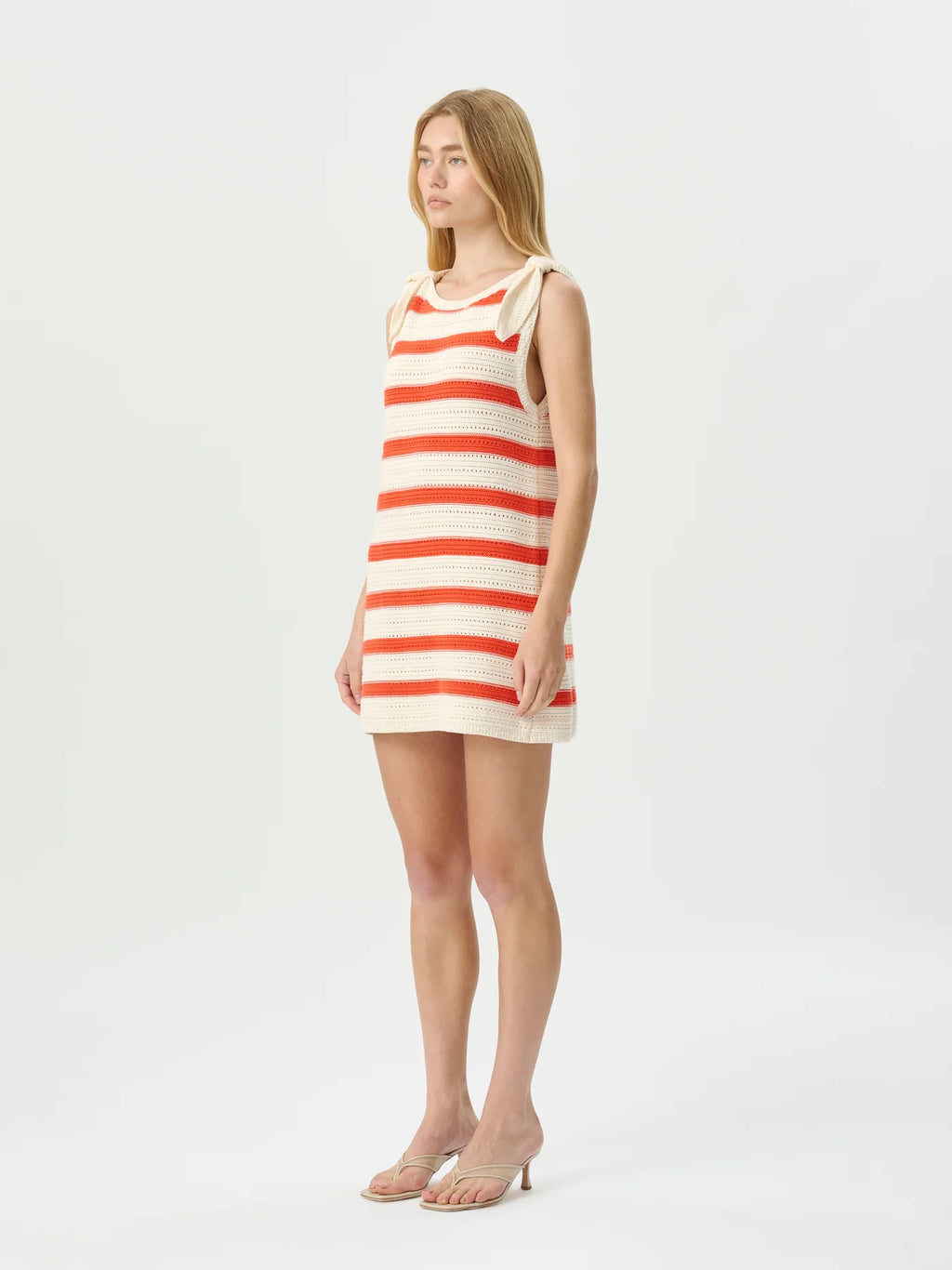 A woman modeling a sleeveless knit mini dress with horizontal red and cream stripes, standing against a plain light gray studio background.