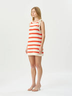 A woman modeling a sleeveless knit mini dress with horizontal red and cream stripes, standing against a plain light gray studio background.