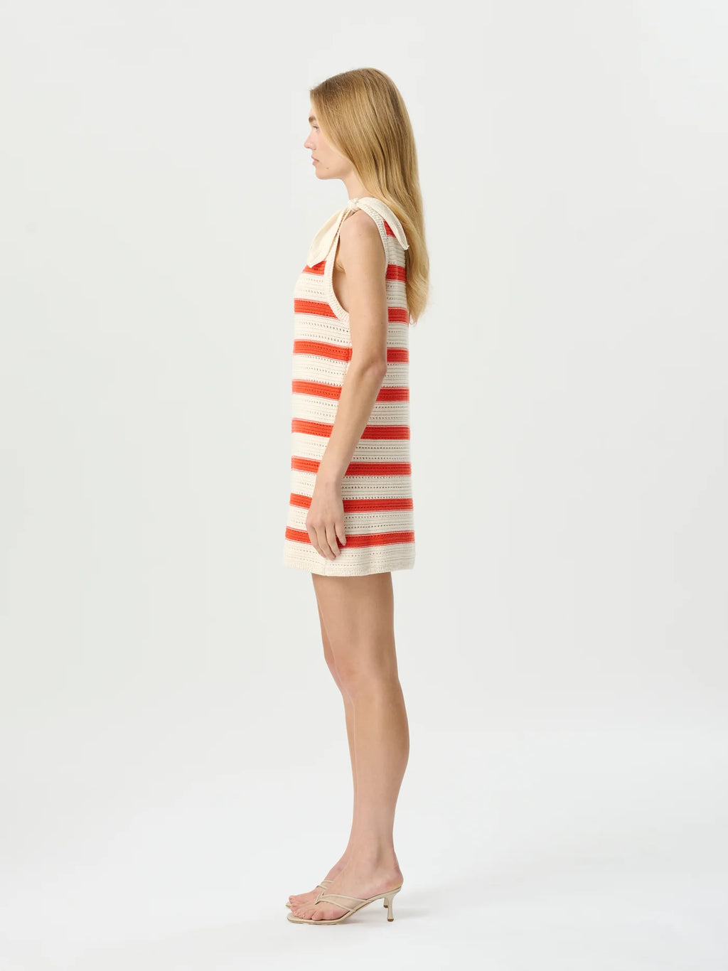 A woman stands in profile wearing a sleeveless cream knit dress with horizontal coral-red stripes and a small bow at the neckline, paired with clear high-heel sandals, against a plain light gray studio background.