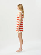 A woman stands in profile wearing a sleeveless cream knit dress with horizontal coral-red stripes and a small bow at the neckline, paired with clear high-heel sandals, against a plain light gray studio background.