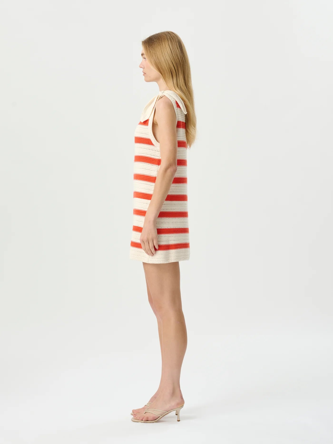 A woman stands in profile wearing a sleeveless cream knit dress with horizontal coral-red stripes and a small bow at the neckline, paired with clear high-heel sandals, against a plain light gray studio background.