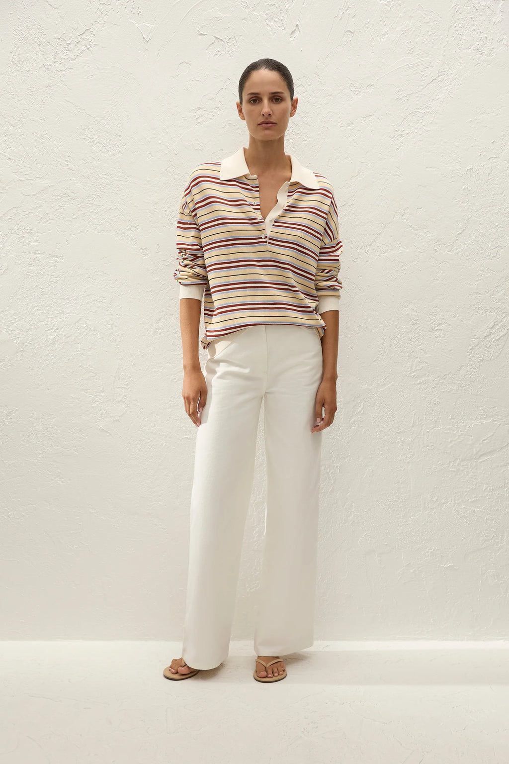 A woman stands against a textured white wall wearing a long-sleeve collared knit top with thin horizontal stripes in beige, brown, and red over a cream base, paired with wide-leg white trousers and tan sandals.
