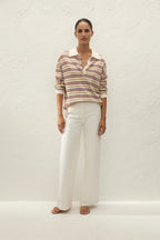 A woman stands against a textured white wall wearing a long-sleeve collared knit top with thin horizontal stripes in beige, brown, and red over a cream base, paired with wide-leg white trousers and tan sandals.
