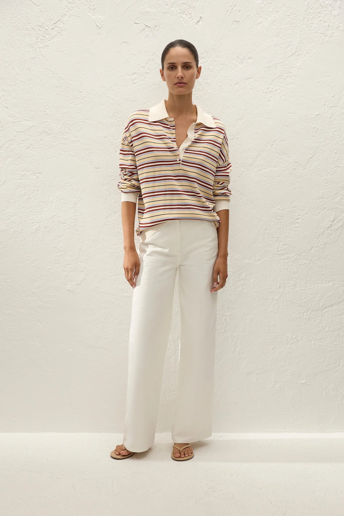 A woman stands against a textured white wall wearing a long-sleeve collared knit top with thin horizontal stripes in beige, brown, and red over a cream base, paired with wide-leg white trousers and tan sandals.