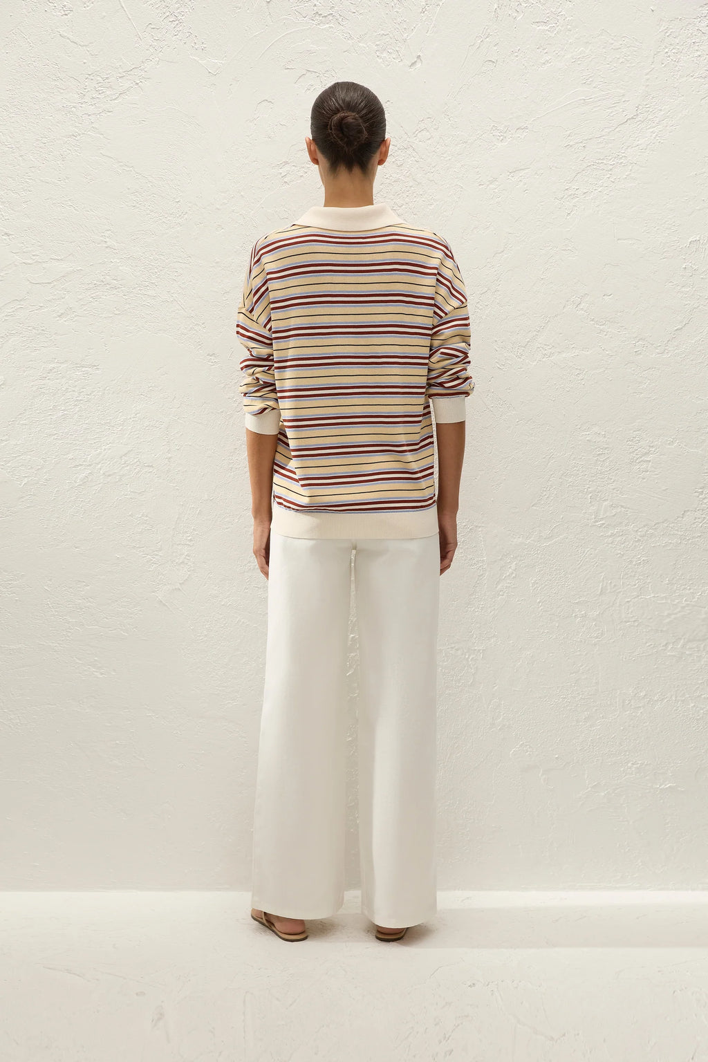 A model viewed from behind wearing a cream-colored long-sleeve knit sweater with thin multicolor horizontal stripes (red, yellow, blue) and white wide-leg trousers, standing against a textured off-white wall.