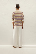 A model viewed from behind wearing a cream-colored long-sleeve knit sweater with thin multicolor horizontal stripes (red, yellow, blue) and white wide-leg trousers, standing against a textured off-white wall.