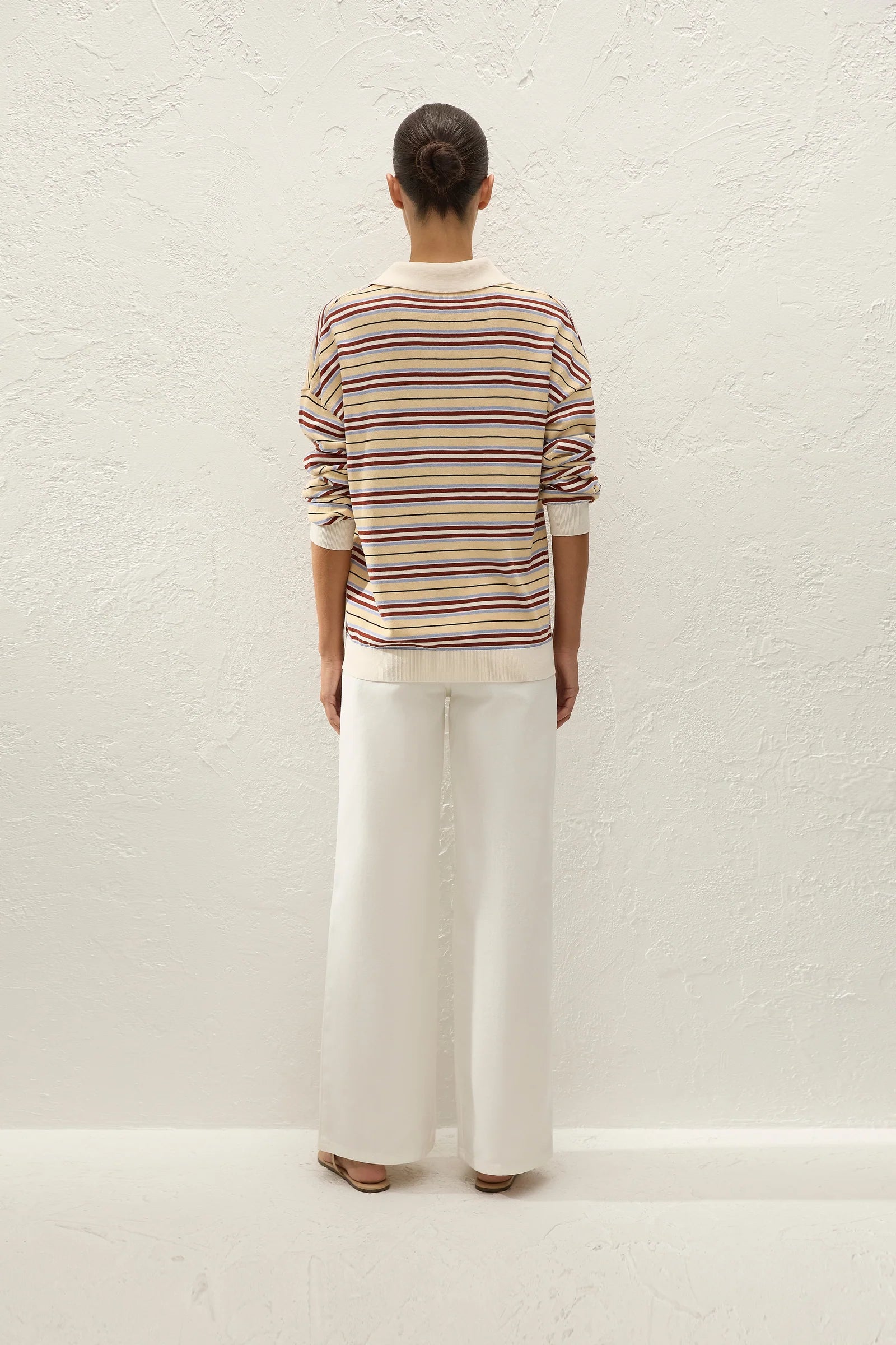 A model viewed from behind wearing a cream-colored long-sleeve knit sweater with thin multicolor horizontal stripes (red, yellow, blue) and white wide-leg trousers, standing against a textured off-white wall.