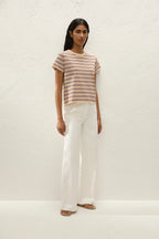 A woman stands against a textured off-white wall wearing a short-sleeve crewneck t-shirt with thin brown and beige horizontal stripes, paired with white wide-leg trousers and flat sandals.