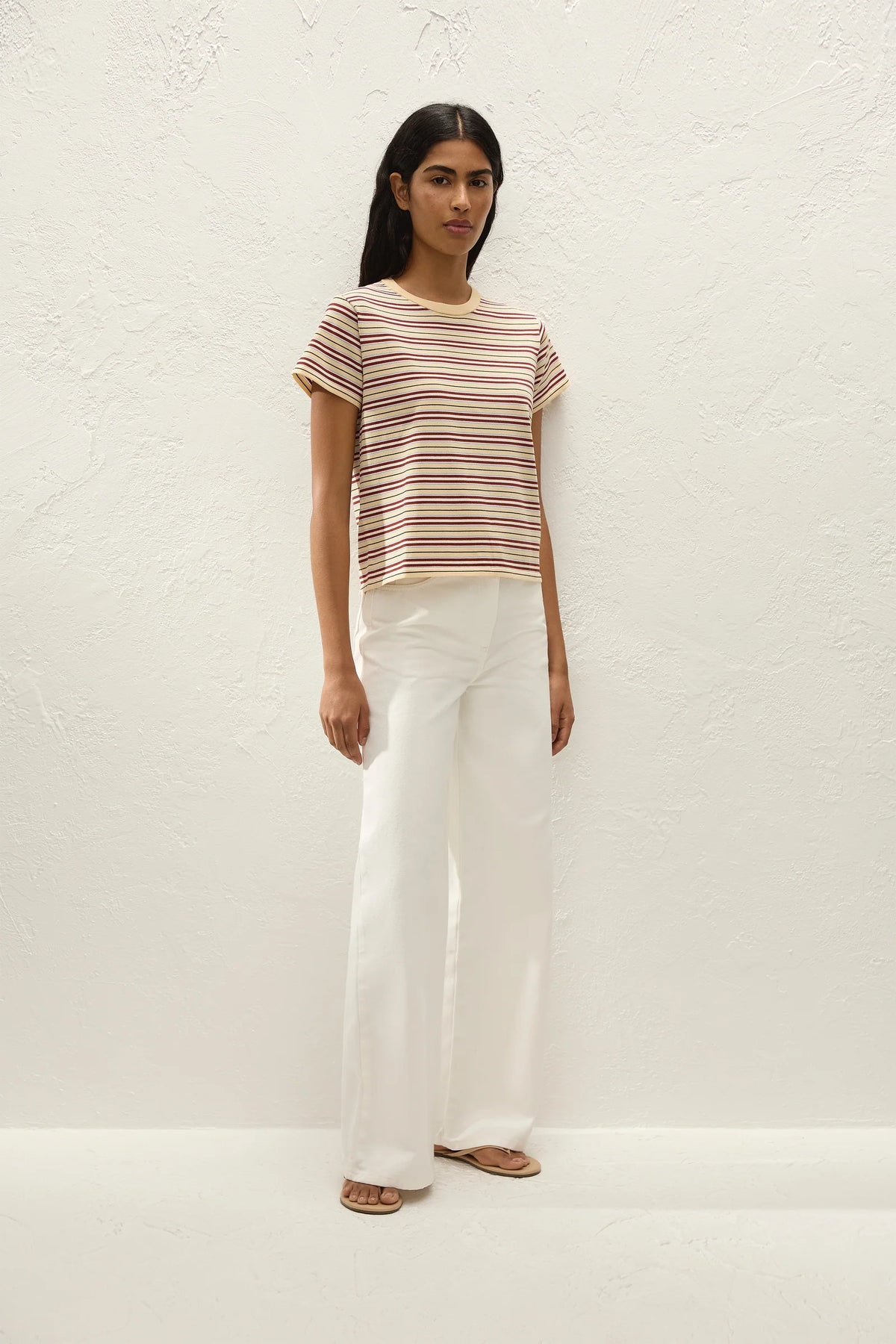 A woman stands against a textured off-white wall wearing a short-sleeve crewneck t-shirt with thin brown and beige horizontal stripes, paired with white wide-leg trousers and flat sandals.