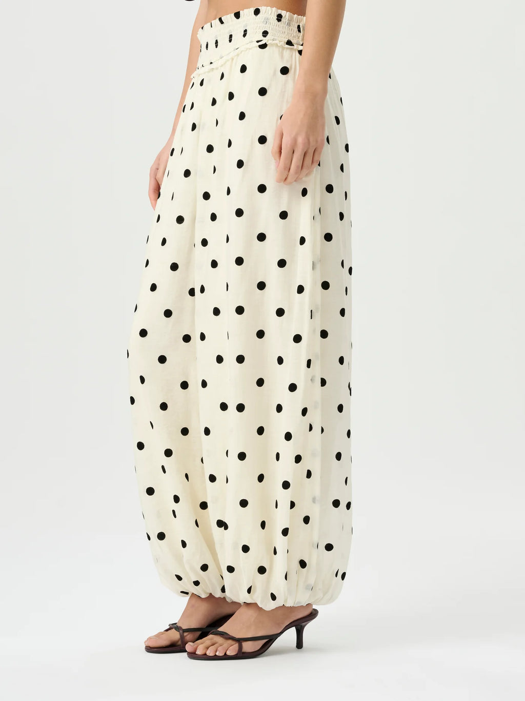 A model wearing high-waisted, voluminous cream-colored pants with a black polka dot pattern, paired with dark brown heeled sandals, against a plain light gray studio background.