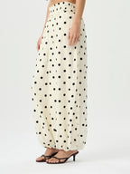 A model wearing high-waisted, voluminous cream-colored pants with a black polka dot pattern, paired with dark brown heeled sandals, against a plain light gray studio background.