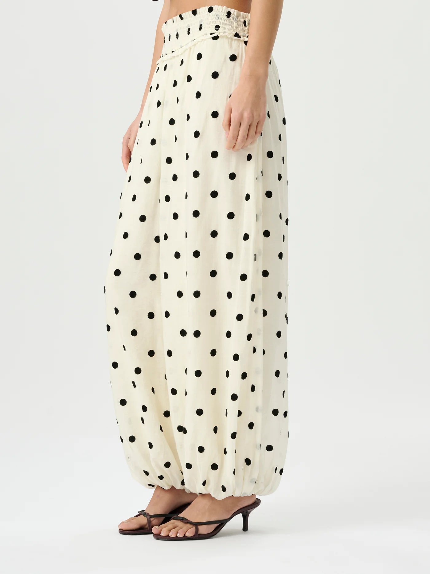A model wearing high-waisted, voluminous cream-colored pants with a black polka dot pattern, paired with dark brown heeled sandals, against a plain light gray studio background.