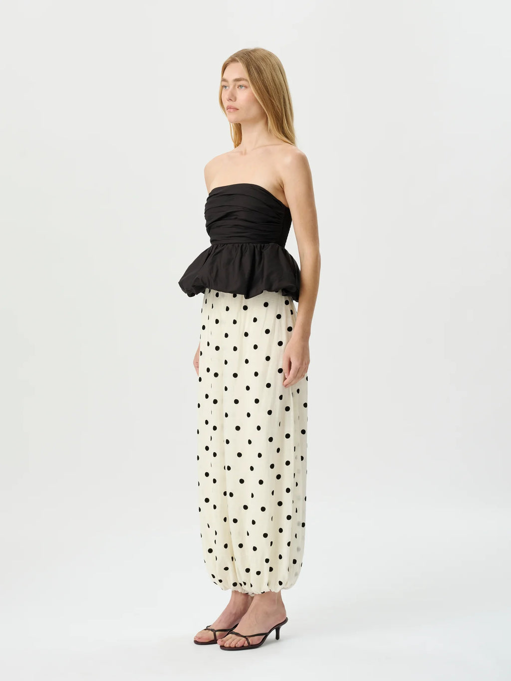 A woman modeling a strapless black peplum top paired with cream wide-leg pants featuring a black polka dot pattern, set against a plain light gray studio background.