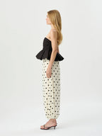 A woman stands in profile wearing a black strapless peplum top and cream wide-leg pants with black polka dots, paired with black heeled sandals, against a plain light gray studio background.