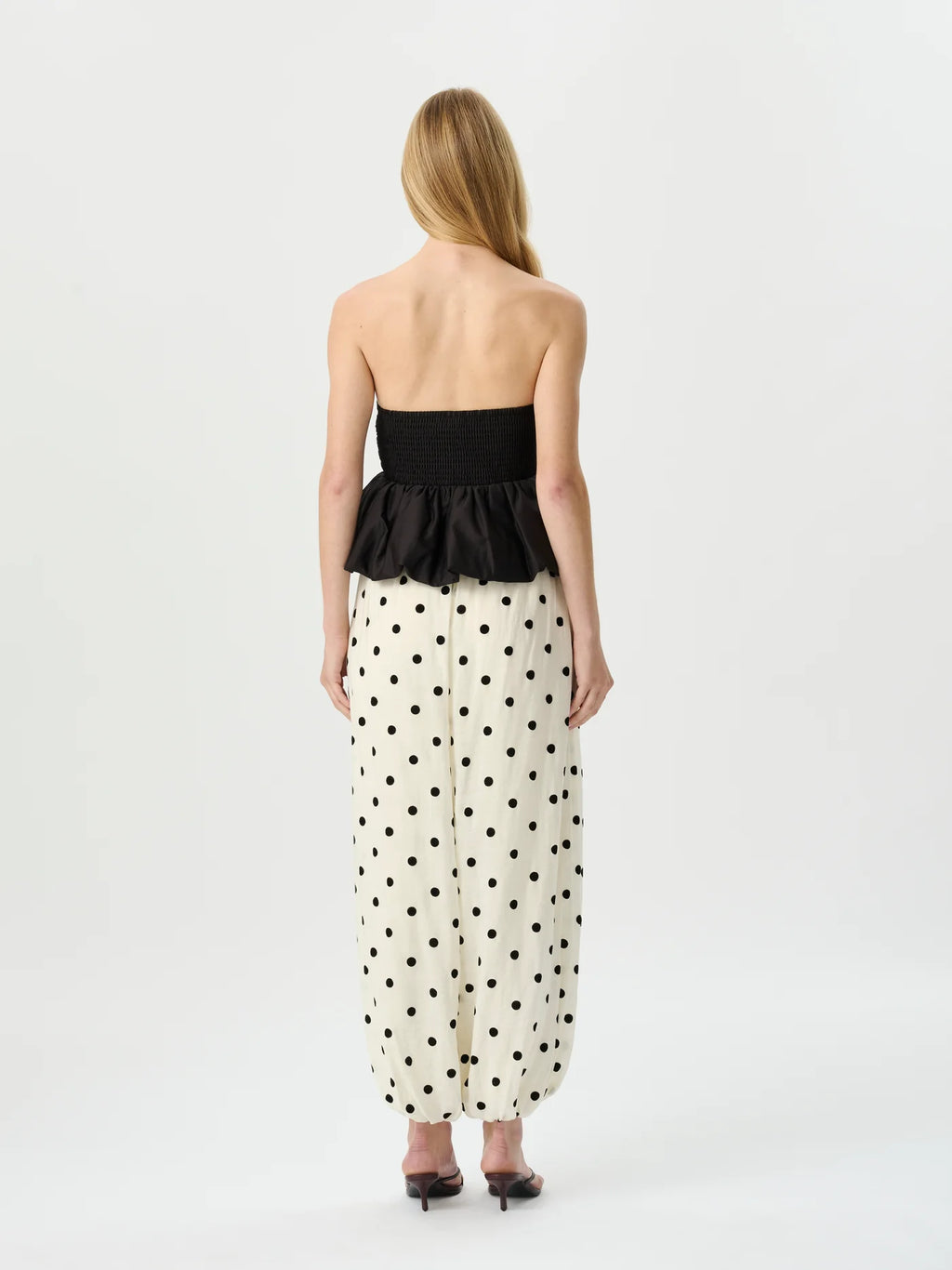 A woman viewed from the back wearing a strapless black peplum top and a cream maxi skirt with black polka dots, set against a plain light gray studio background.