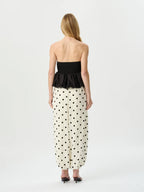 A woman viewed from the back wearing a strapless black peplum top and a cream maxi skirt with black polka dots, set against a plain light gray studio background.