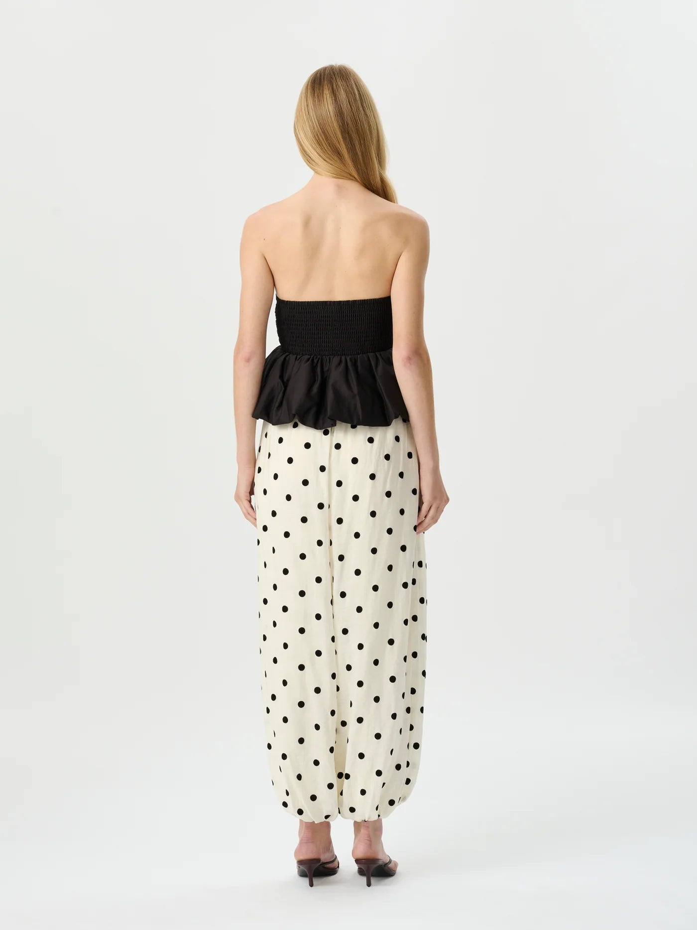 A woman viewed from the back wearing a strapless black peplum top and a cream maxi skirt with black polka dots, set against a plain light gray studio background.