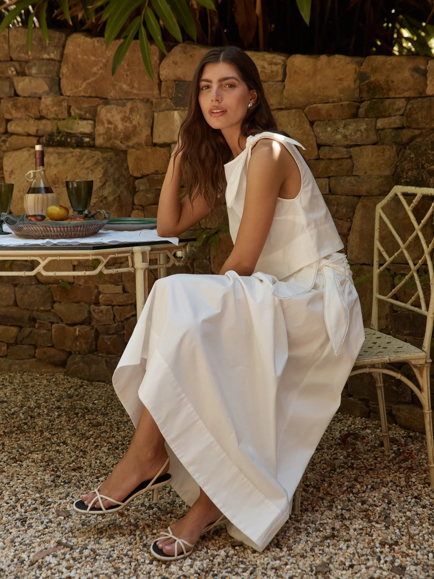 Woman in a white dress sitting outdoors near a stone wall.