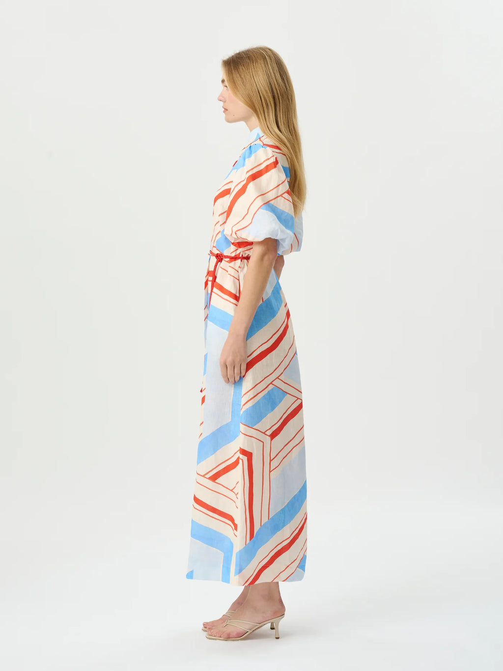 A woman stands in profile wearing a long wrap-style maxi dress with a geometric stripe pattern in light blue, coral red, and cream on a white base, paired with nude high-heeled sandals, against a plain white studio background.