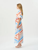 A woman stands in profile wearing a long wrap-style maxi dress with a geometric stripe pattern in light blue, coral red, and cream on a white base, paired with nude high-heeled sandals, against a plain white studio background.