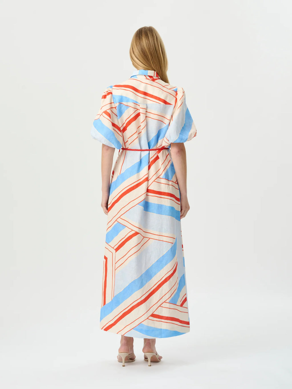 A woman viewed from the back wearing a short-sleeve maxi dress with a white base and diagonal stripes in light blue and orange-red, paired with beige high heels against a plain white studio background.