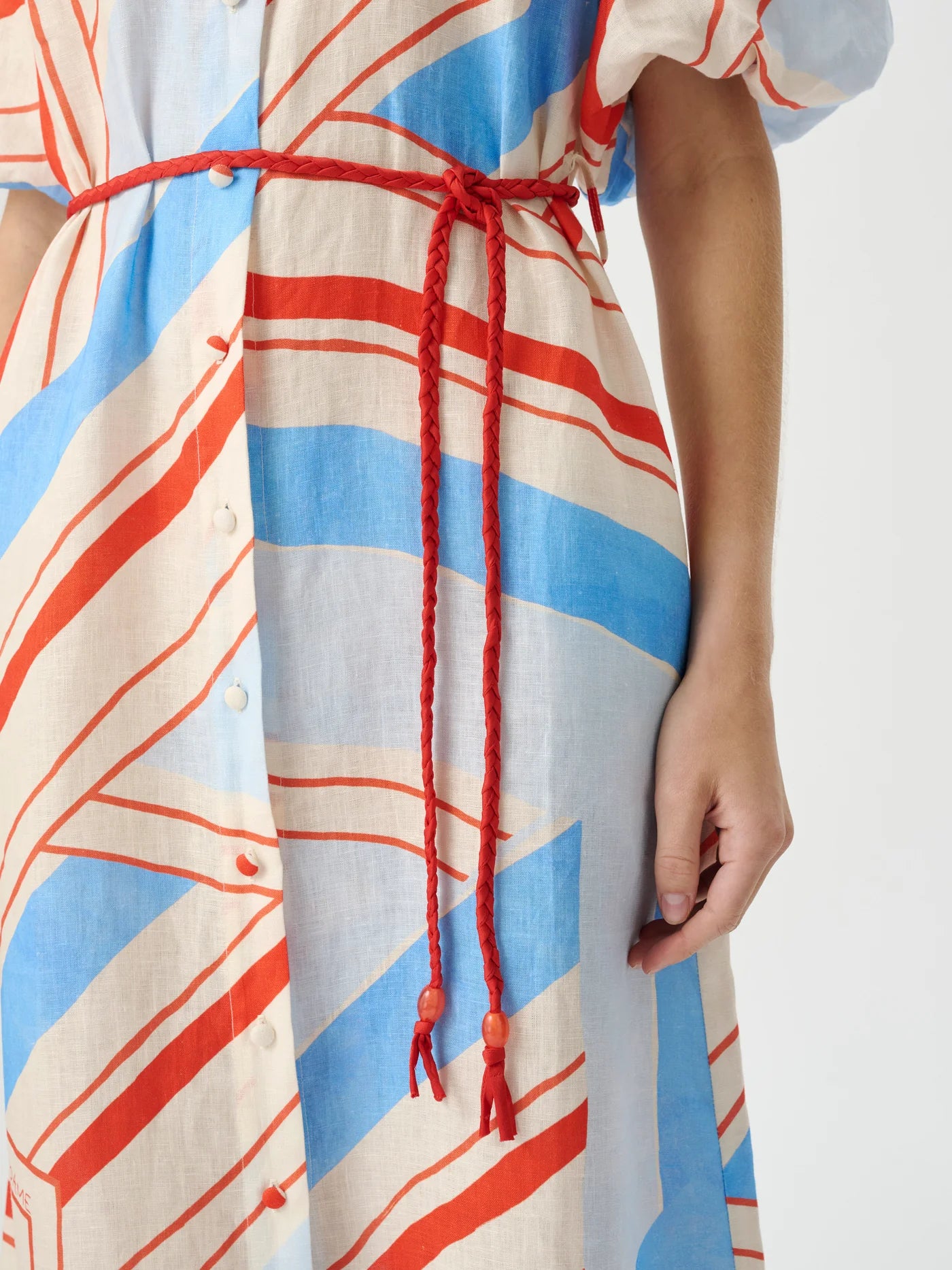 A close-up of a person wearing a loose dress with a blue, orange, and cream abstract stripe pattern, featuring a red braided drawstring belt tied at the waist.