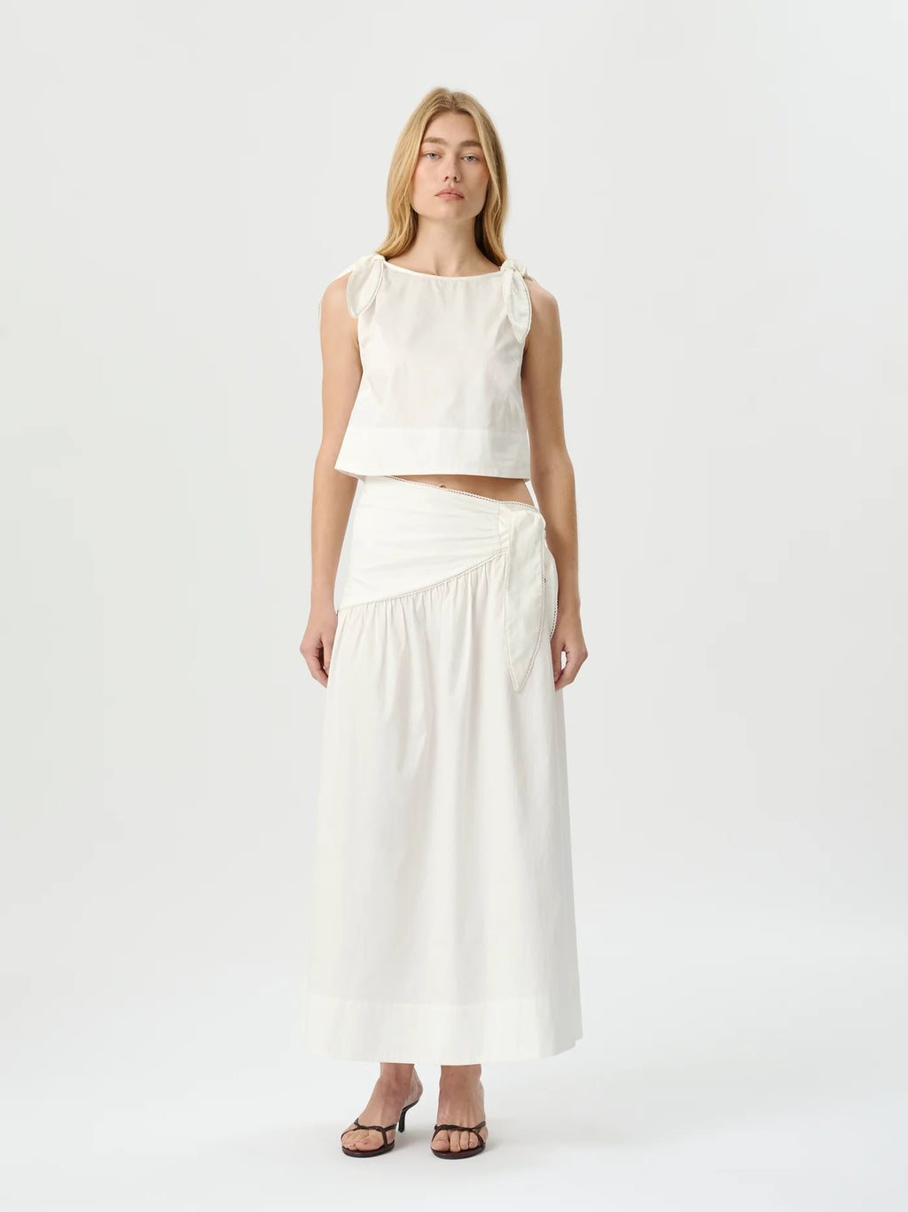 A woman modeling a white sleeveless crop top and matching high-waisted wrap maxi skirt against a plain white studio background.
