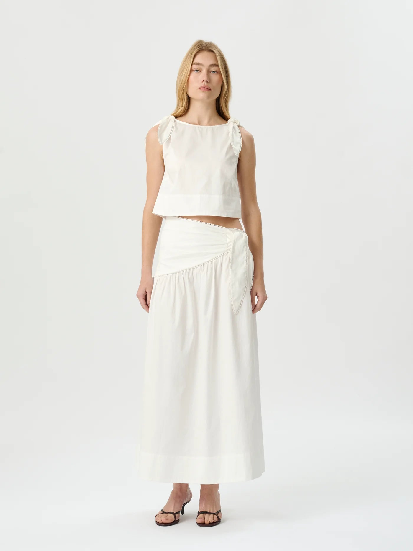 A woman modeling a white sleeveless crop top and matching high-waisted wrap maxi skirt against a plain white studio background.