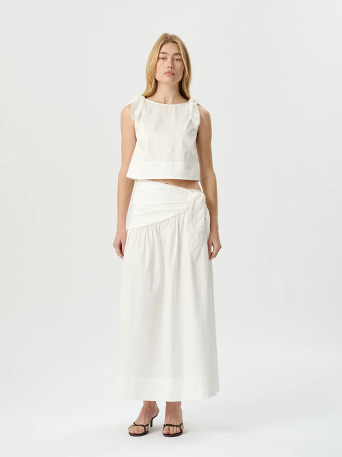 A woman modeling a white sleeveless crop top and matching high-waisted wrap maxi skirt against a plain white studio background.