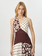 A woman modeling a halter-neck dress with a patchwork design of cream, brown, and burgundy floral and polka-dot fabrics against a light gray studio background.